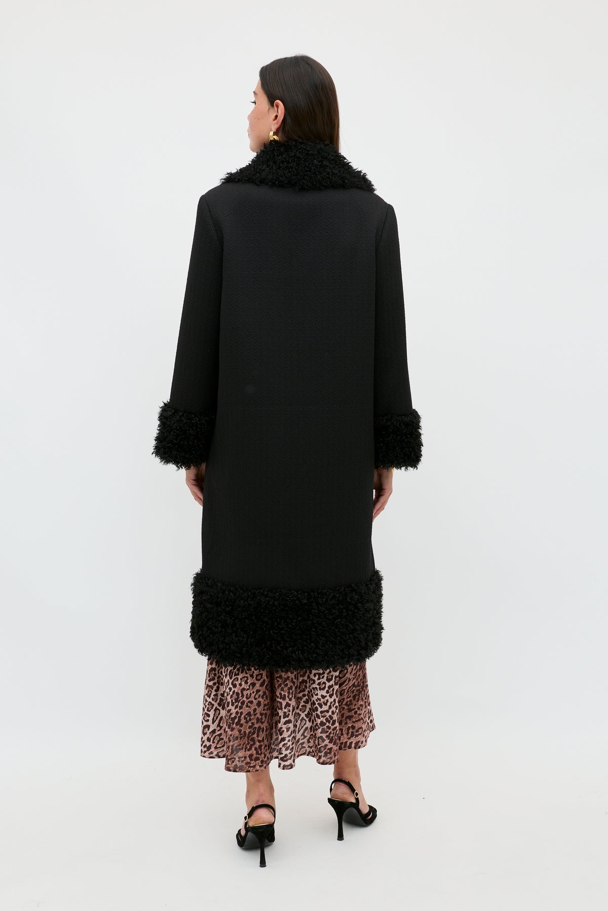 A person stands facing away, wearing the Diara - Black—a long black coat with faux-fur shearling trim on the cuffs and hem—paired with a leopard print skirt and black heels, set against a plain white background.