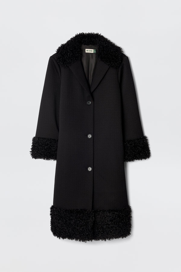 The Diara - Black is a long, black 70s-style coat with three buttons and fluffy faux-fur shearling trim on the collar, cuffs, and hem, shown against a plain white background.