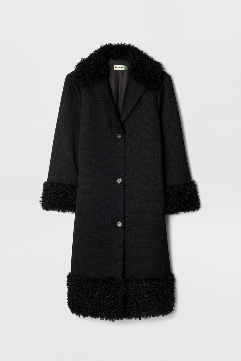 The Diara - Black is a long, black 70s-style coat with three buttons and fluffy faux-fur shearling trim on the collar, cuffs, and hem, shown against a plain white background.