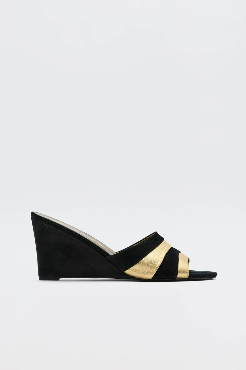 The Alira - Gold is a '70s-inspired black and gold wedge sandal with an open toe and wide crisscross straps, featuring bold gold accents. Suede-like wedge heel and straps contrast against a plain white background.