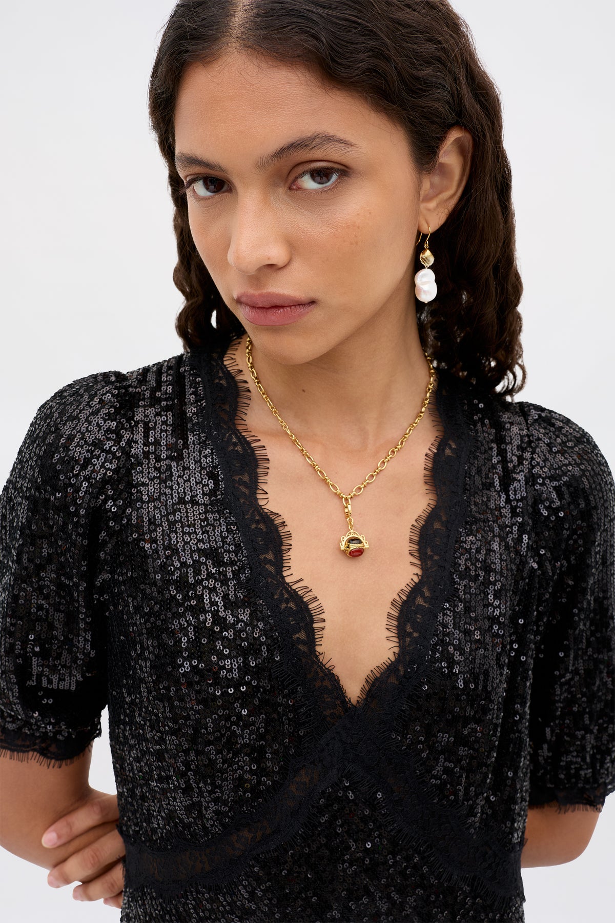 A woman with wavy dark hair wears a black sequin top, the Sariah - Gold necklace featuring a red semi-precious stone pendant, and dangling pearl earrings that evoke vintage charm. She looks directly at the camera with a neutral expression against a plain background.
