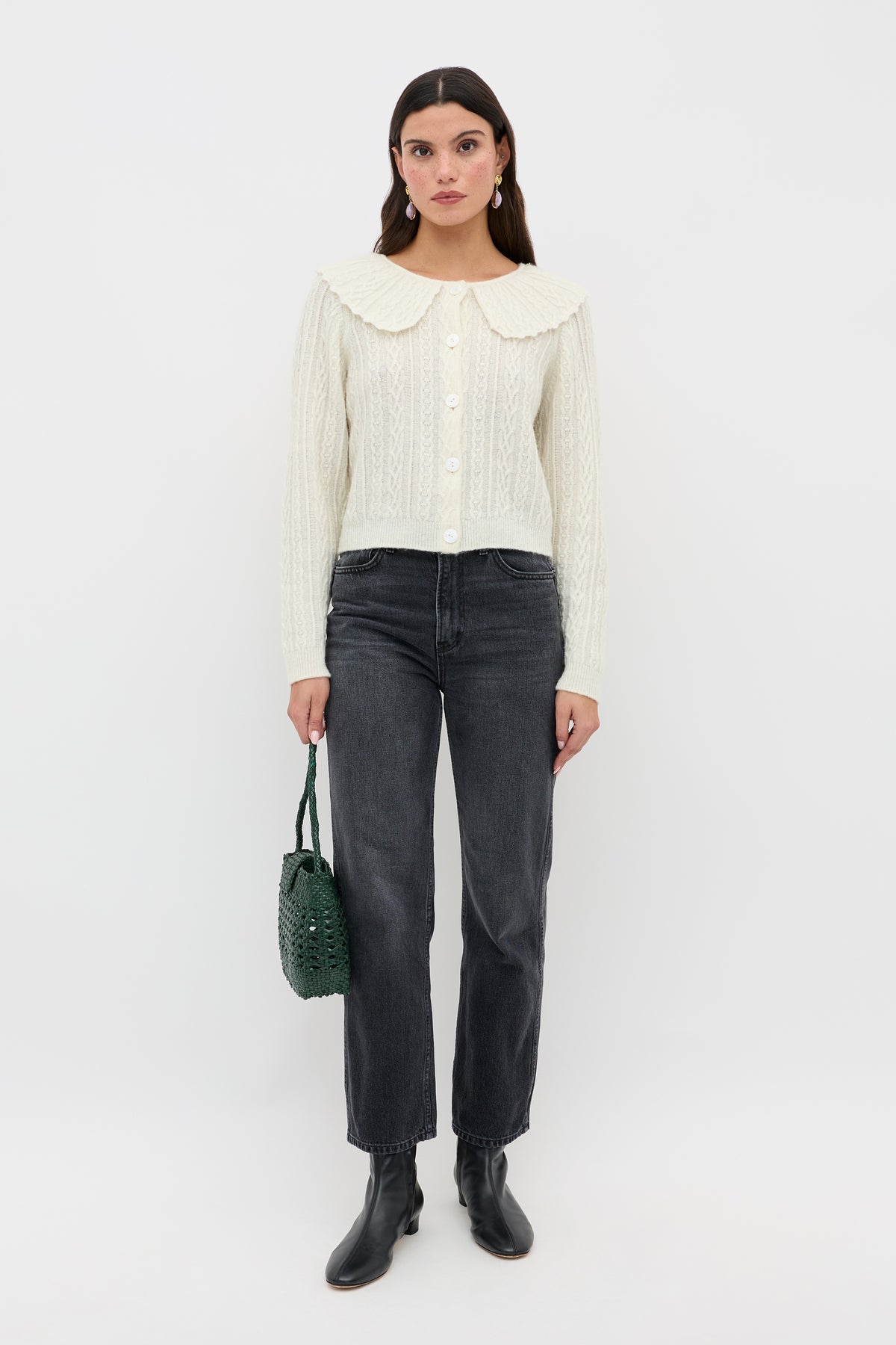 A woman stands against a plain white background wearing the Jelena - Cream cable knit cardigan with a large collar, paired with black jeans, black ankle boots, and holding a green textured handbag.