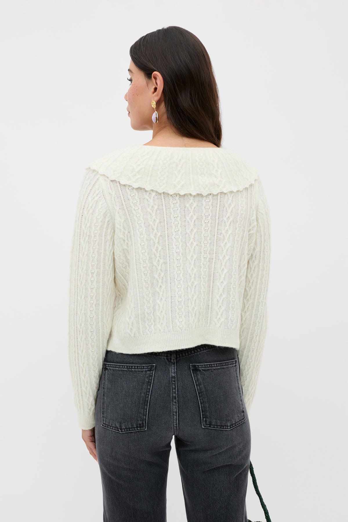 A person wearing the Jelena - Cream cable knit cardigan, featuring a wide collar, paired with dark high-waisted jeans stands facing away against a plain white background.