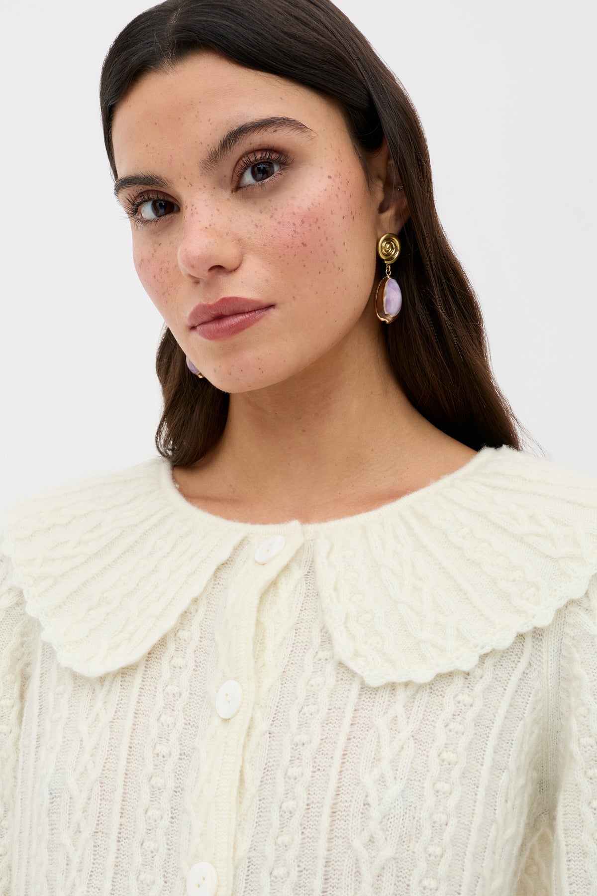 A woman with long brown hair and freckles wears the Jelena - Cream cable knit cardigan, featuring a large scalloped collar, paired with pearl earrings. She gazes slightly to the side against a plain light background.