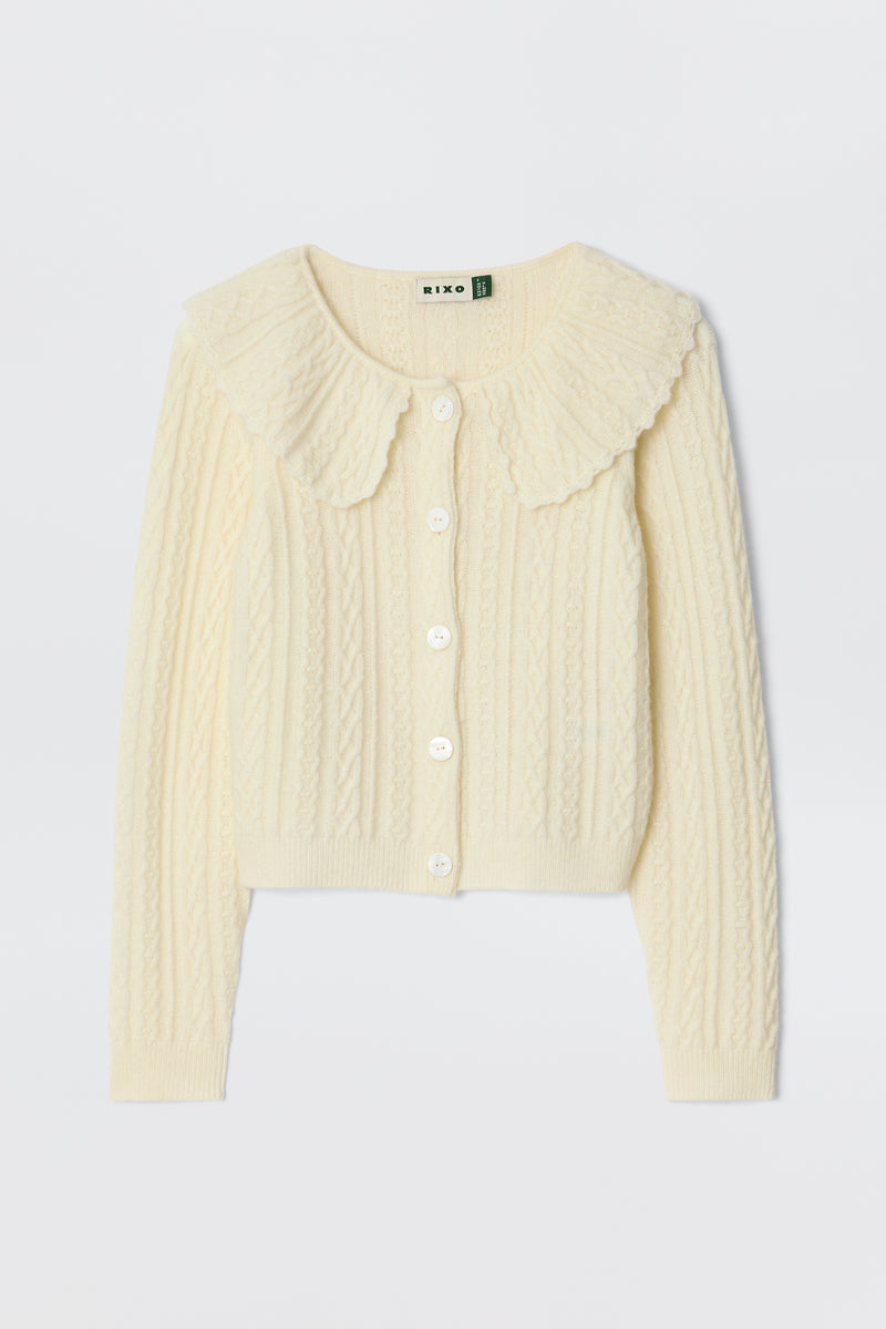 The Jelena - Cream is a cream-colored, long-sleeve cropped cable knit cardigan with white buttons and a large scalloped collar—ideal for layering between seasons. Laid flat on a white background.
