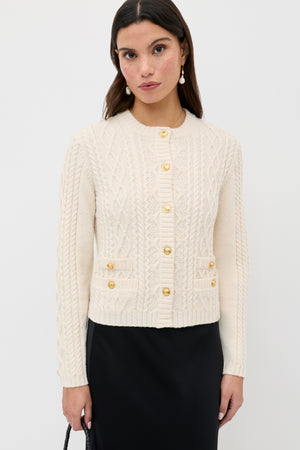 A woman wearing the Kamina - Cream cable-knit cardigan with gold buttons and a black skirt stands against a plain white background. She has long dark hair and accessorizes with drop earrings.