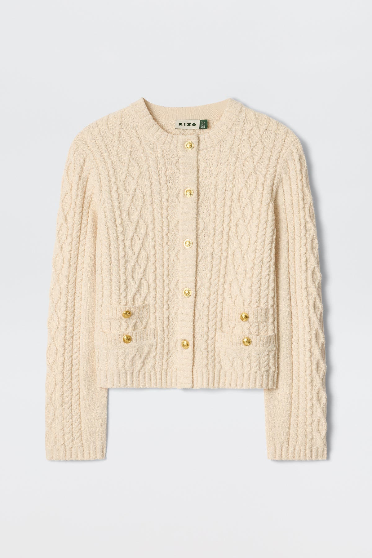 The Kamina - Cream is a cream-colored, long-sleeve cable knit cardigan featuring gold buttons and two front pockets, displayed on a plain white background.