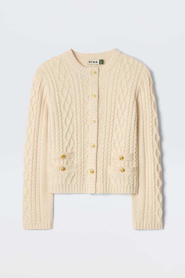 The Kamina - Cream is a cream-colored, long-sleeve cable knit cardigan featuring gold buttons and two front pockets, displayed on a plain white background.