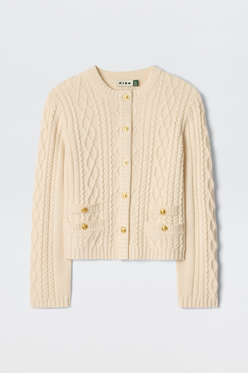 The Kamina - Cream is a cream-colored, long-sleeve cable knit cardigan featuring gold buttons and two front pockets, displayed on a plain white background.