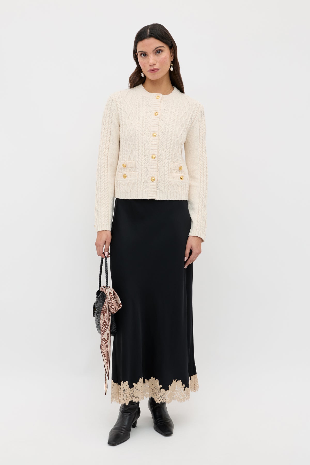 A woman wearing the Kamina - Cream cable knit cardigan with gold buttons, styled with a long black skirt featuring beige lace trim, black ankle boots, and holding a black handbag adorned with a patterned scarf, stands against a white background.