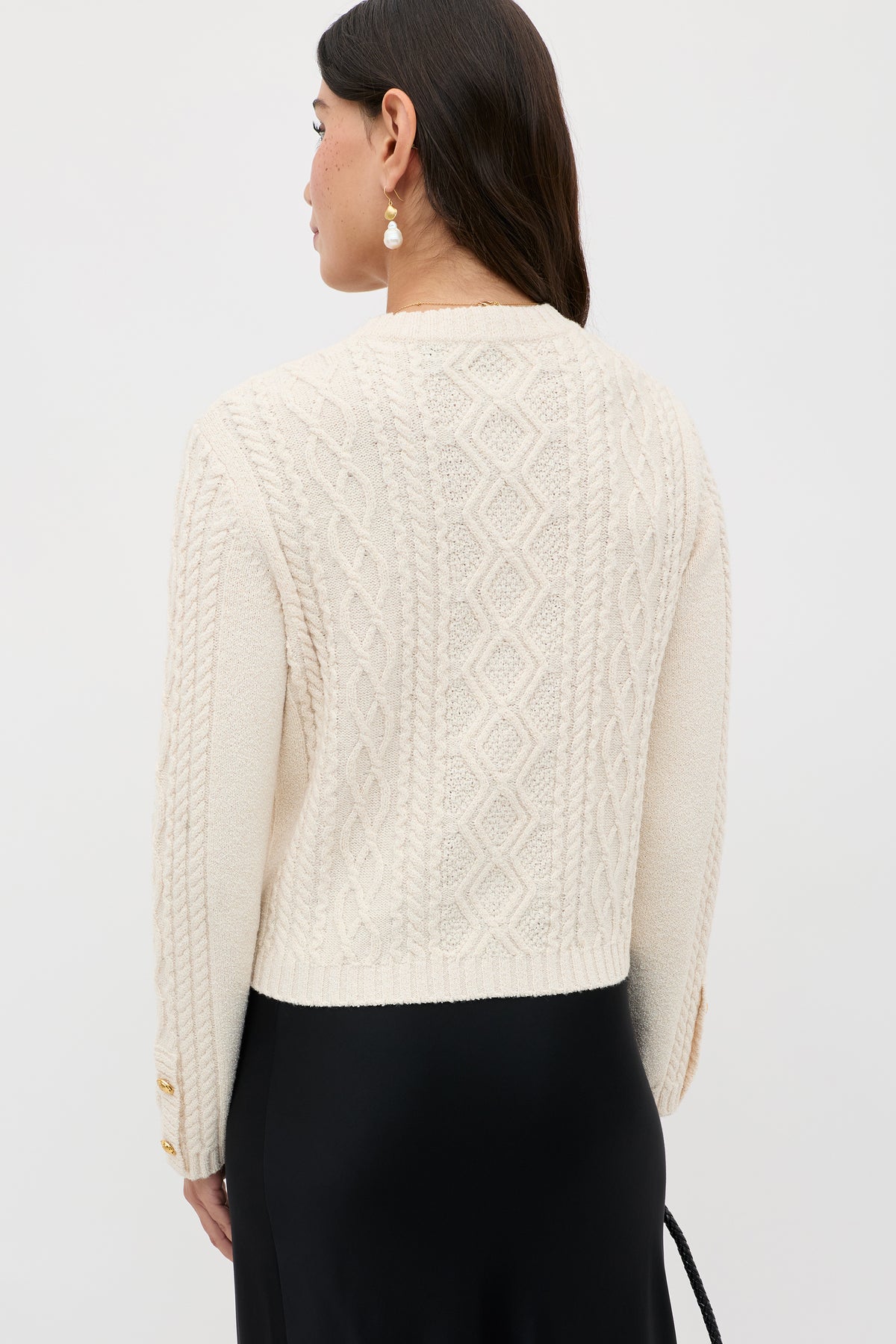 A woman with long dark hair stands with her back to the camera, wearing the Kamina – Cream sweater featuring cable-knit diamond and braid patterns. She pairs it with a black skirt and pearl drop earrings.