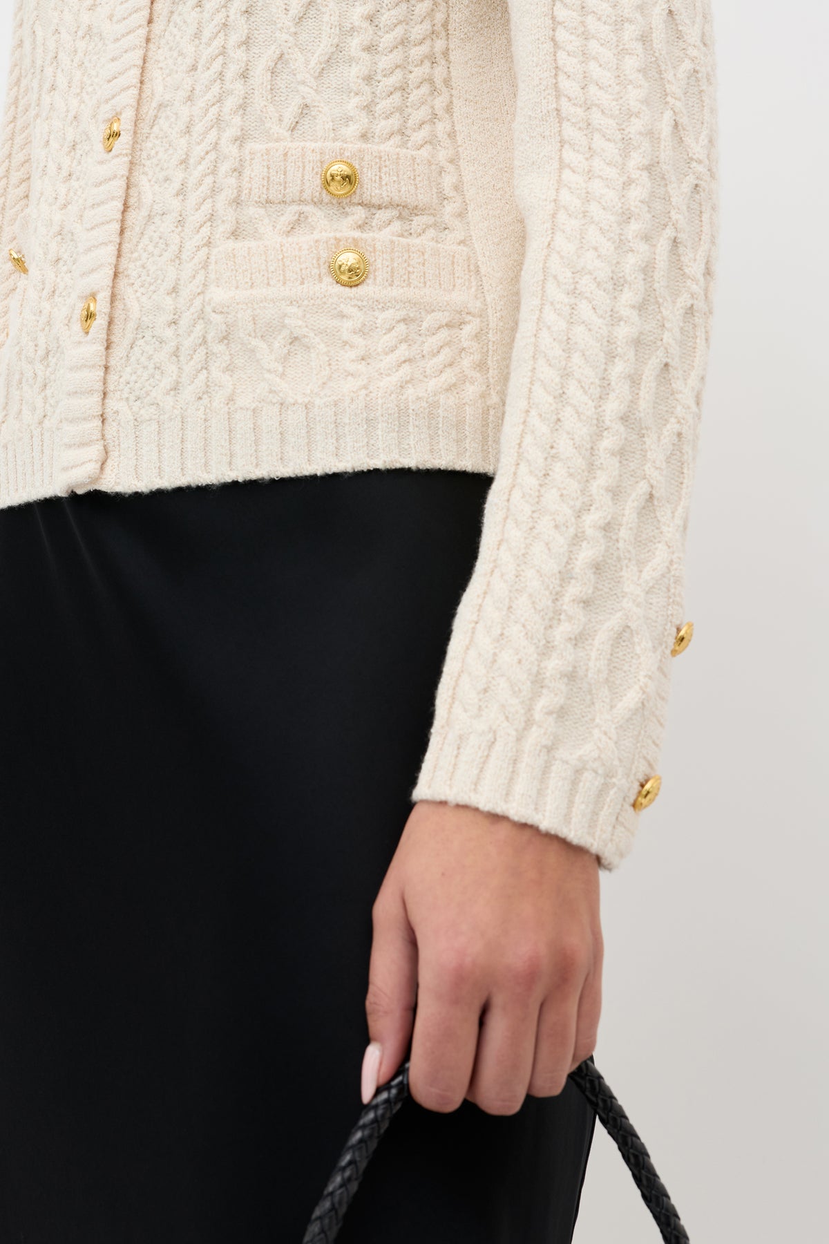 Wearing the Kamina - Cream cable-knit sweater with gold buttons, paired with a black skirt and holding a bag with a black braided handle.