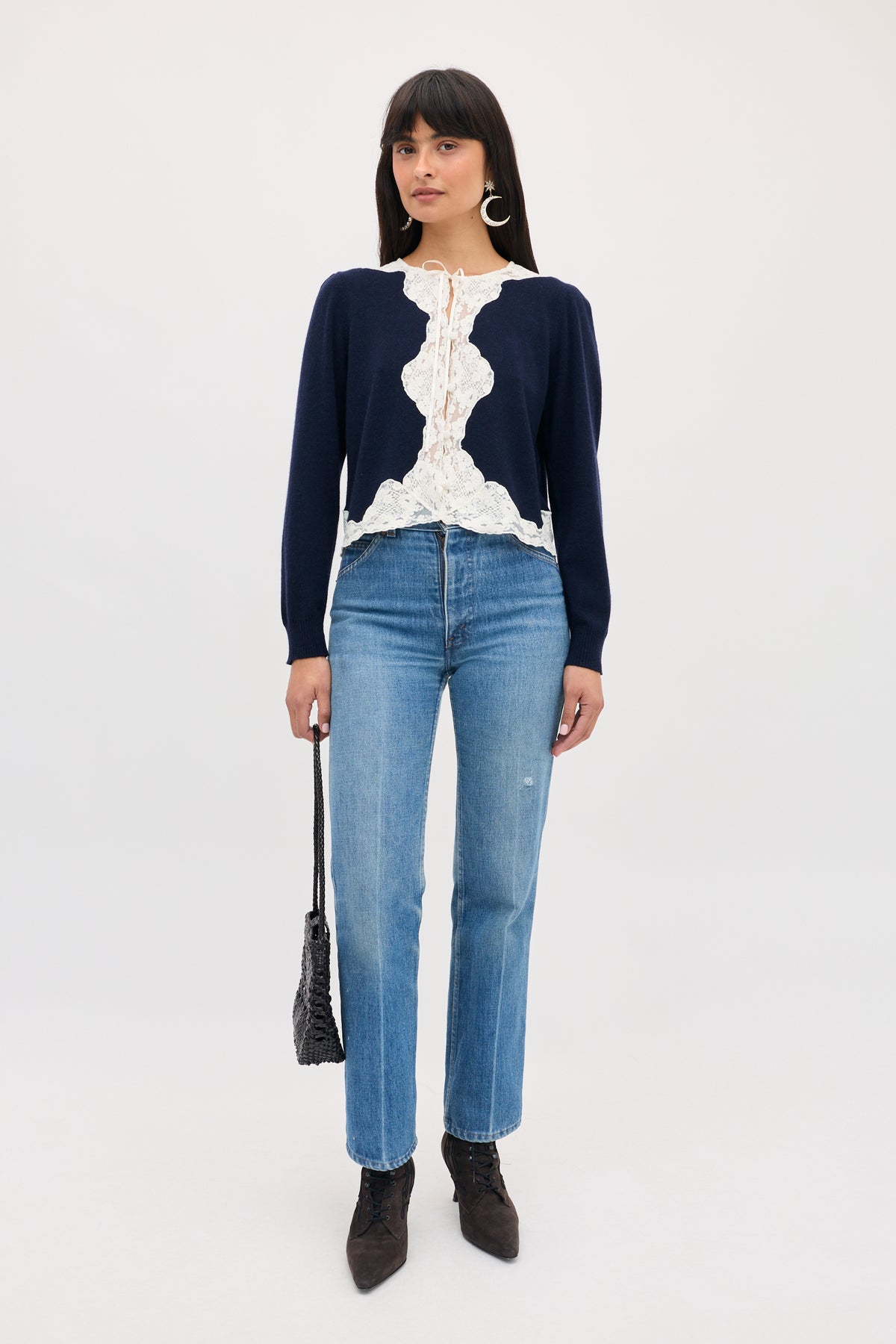A woman wearing a Tassia - Navy long-sleeve cardigan with white lace trim, light-wash blue jeans, and dark boots, carrying a small black woven handbag.