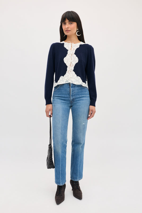 A woman wearing a Tassia - Navy long-sleeve cardigan with white lace trim, light-wash blue jeans, and dark boots, carrying a small black woven handbag.