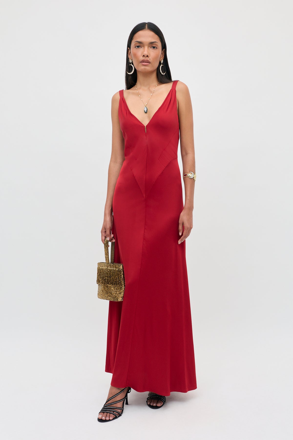 A woman in the “Adelina - Red” sleeveless gown with a V-neckline, holding a textured gold handbag, wearing black strappy heels, hoop earrings, and layered necklaces, stands against a plain light background.