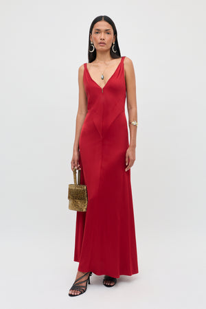 A woman in the “Adelina - Red” sleeveless gown with a V-neckline, holding a textured gold handbag, wearing black strappy heels, hoop earrings, and layered necklaces, stands against a plain light background.