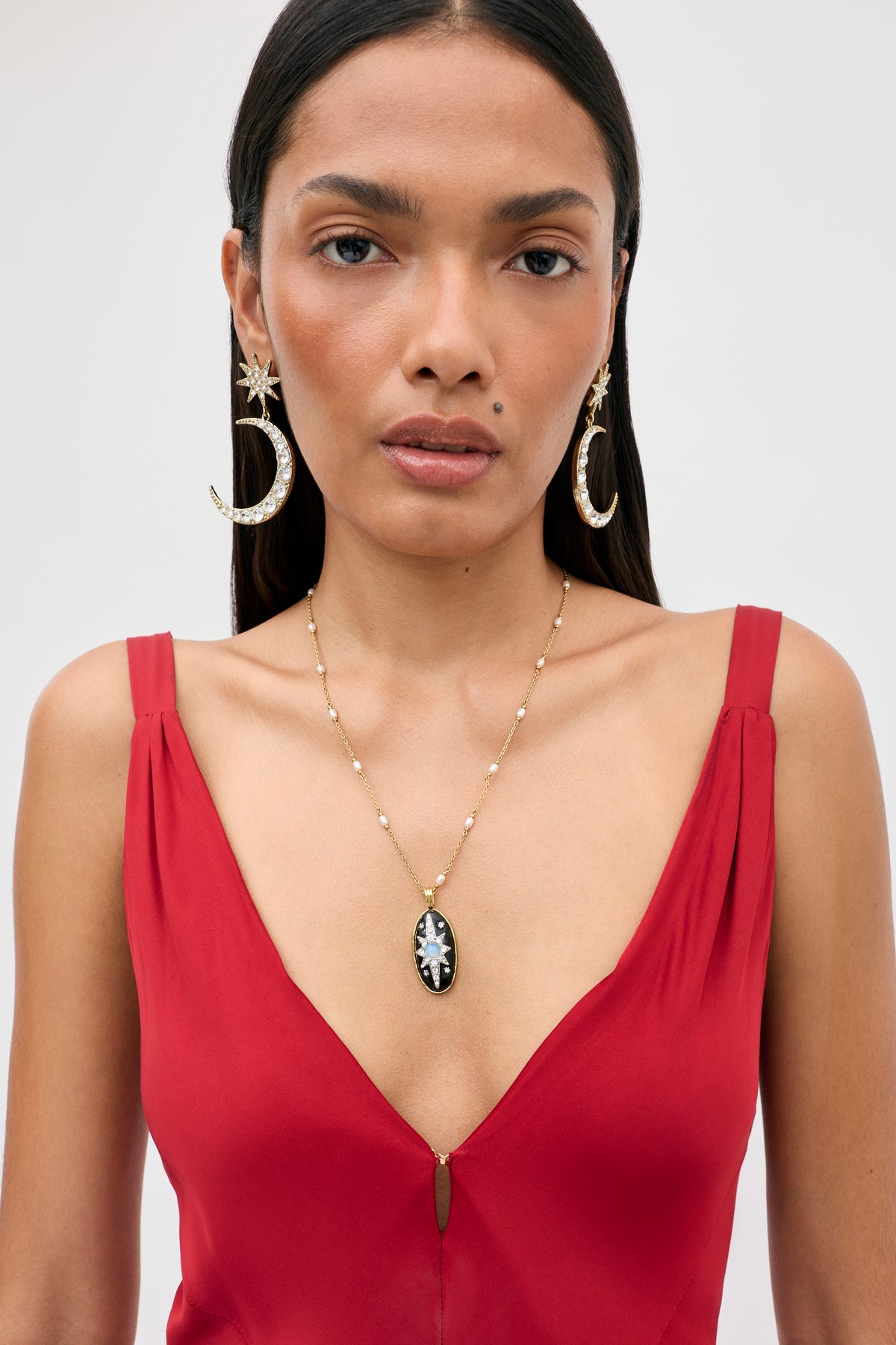 Wearing the “Adelina - Red” dress, the woman pairs her outfit with large crescent moon and star earrings and a gold necklace featuring a black pendant. Her straight dark hair frames her neutral expression as she stands against a plain background.