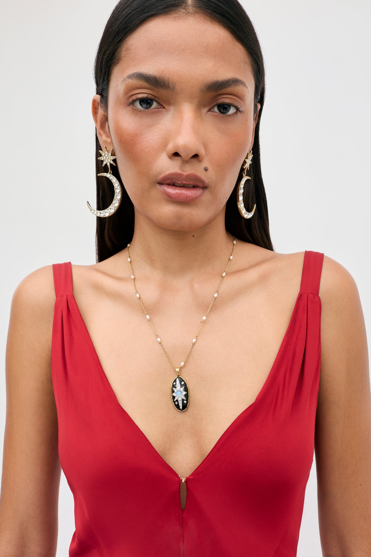 Wearing the “Adelina - Red” dress, the woman pairs her outfit with large crescent moon and star earrings and a gold necklace featuring a black pendant. Her straight dark hair frames her neutral expression as she stands against a plain background.