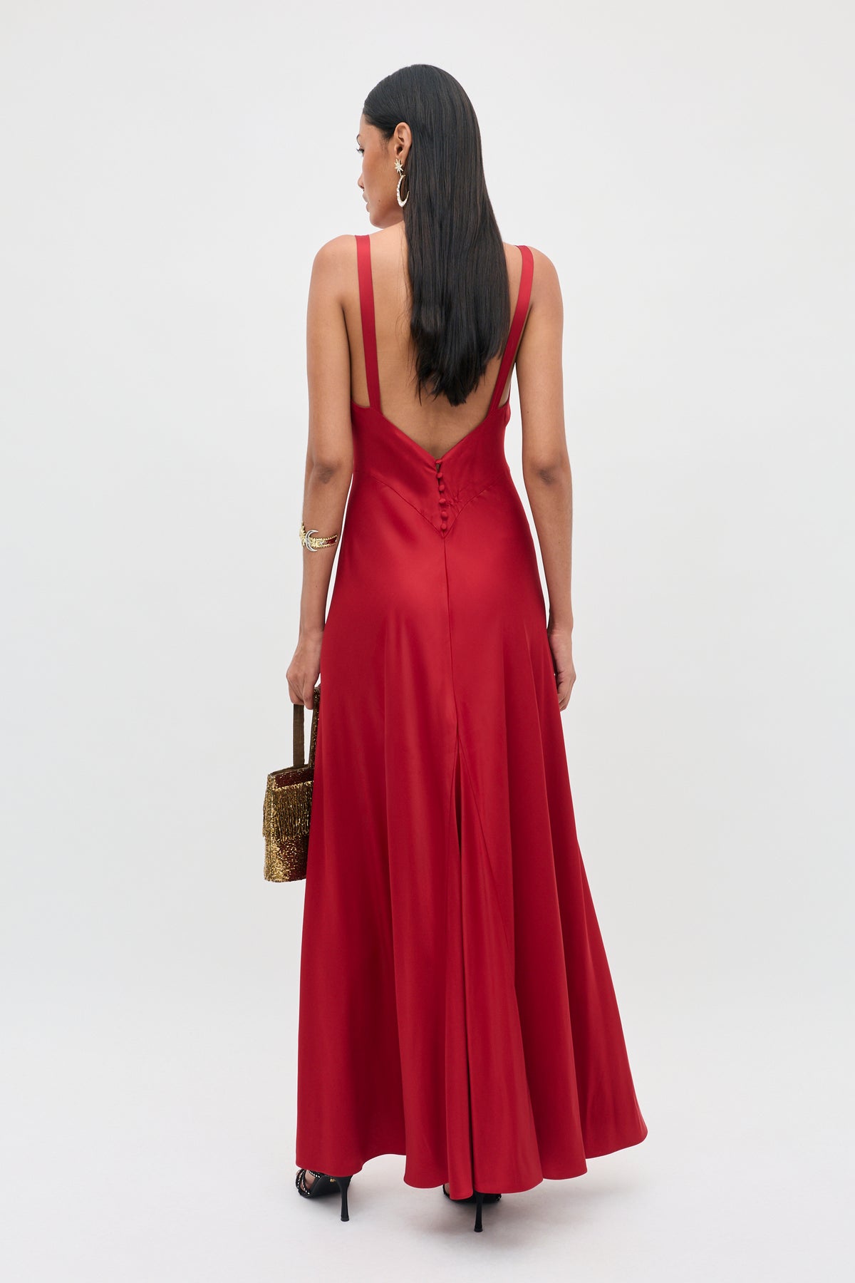 A woman stands with her back to the camera wearing the Adelina - Red sleeveless floor-length gown with a low back and a small row of buttons, holding a small gold handbag.