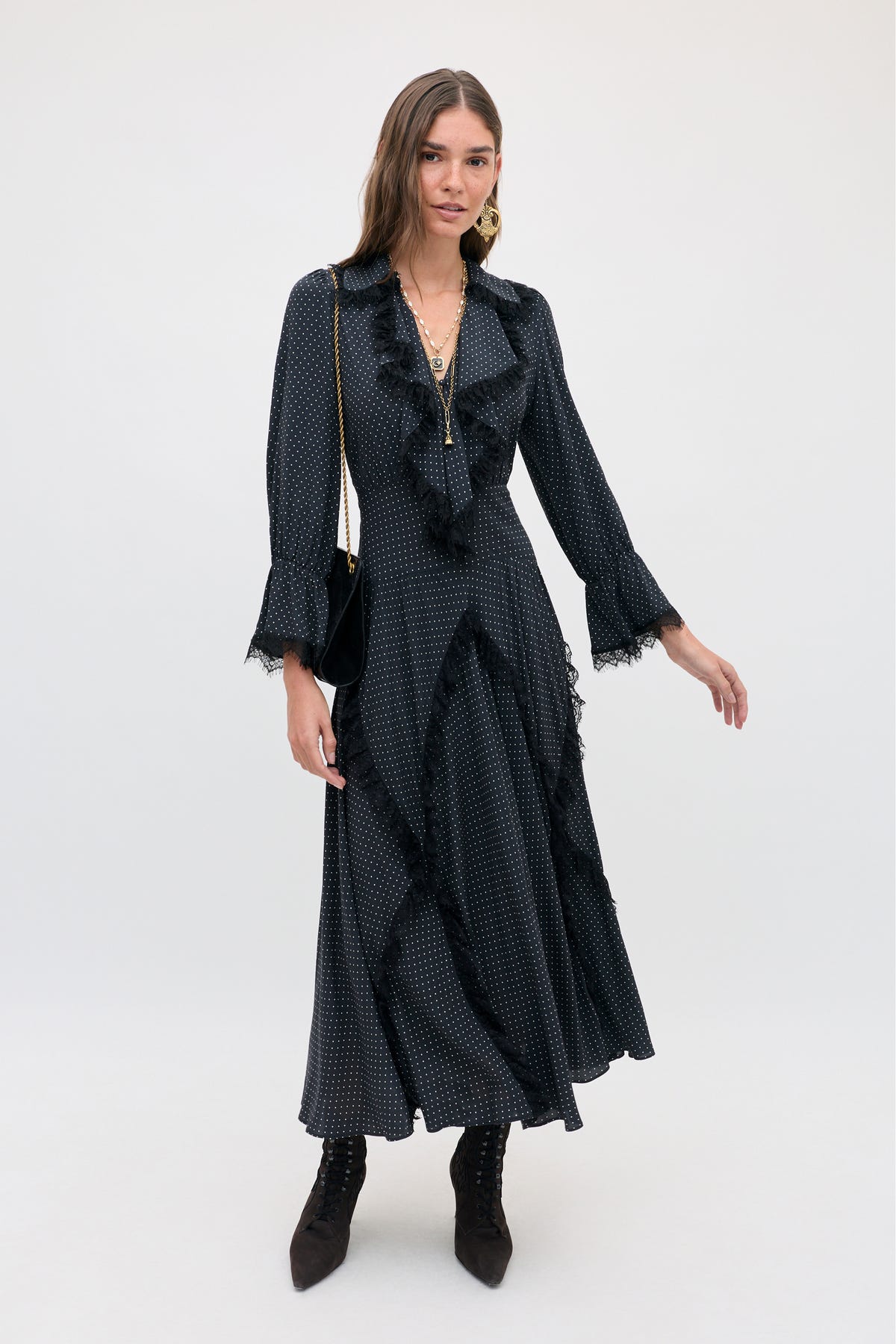 A woman wearing the Aerin - Micro Polka Dot Black midi-length dress with white micro polka dots, black lace trim, and a ruffled neckline, paired with black lace-up ankle boots.