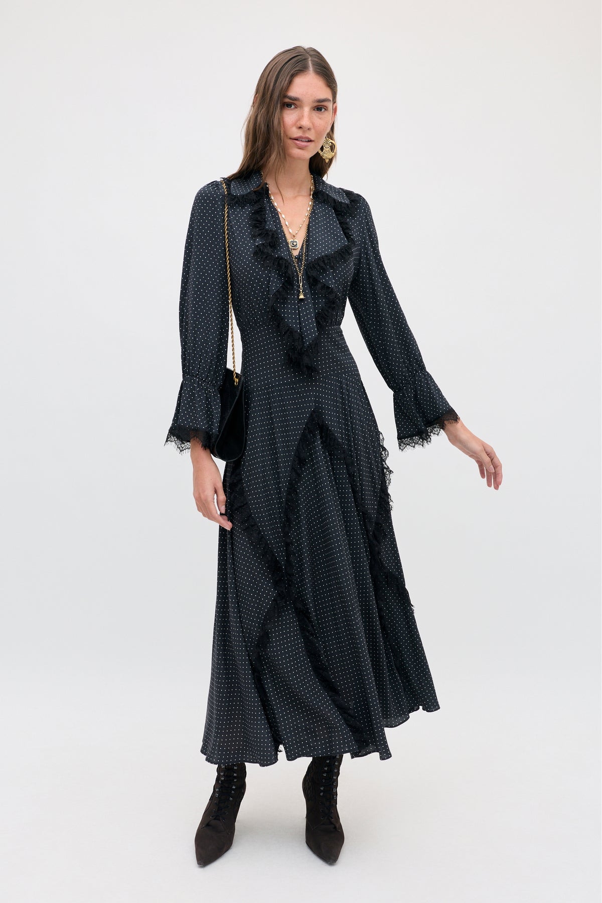 A woman wearing the Aerin - Micro Polka Dot Black midi-length dress with white micro polka dots, black lace trim, and a ruffled neckline, paired with black lace-up ankle boots.