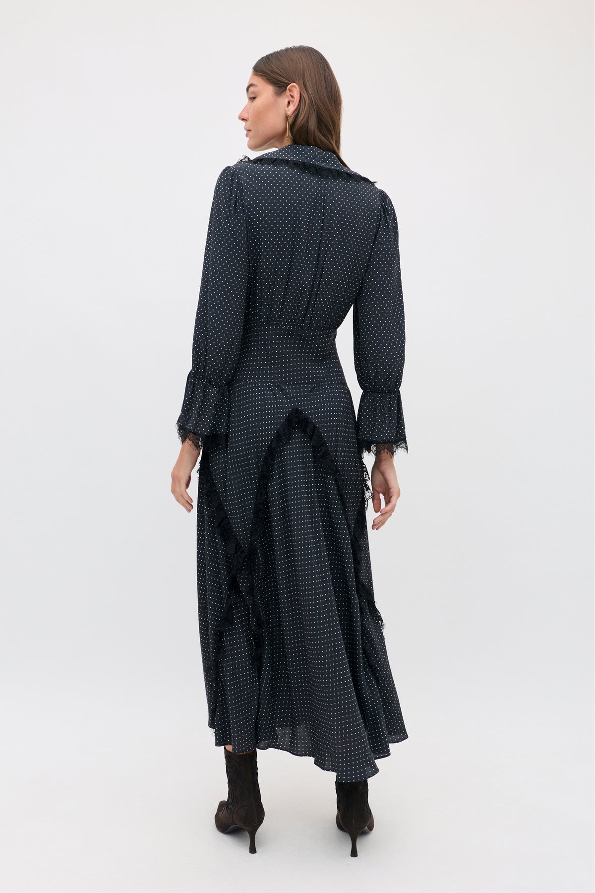 A woman stands facing away wearing the Aerin - Micro Polka Dot Black, a long-sleeved, floor-length black dress with white polka dots, black lace trim, and dark ankle boots.