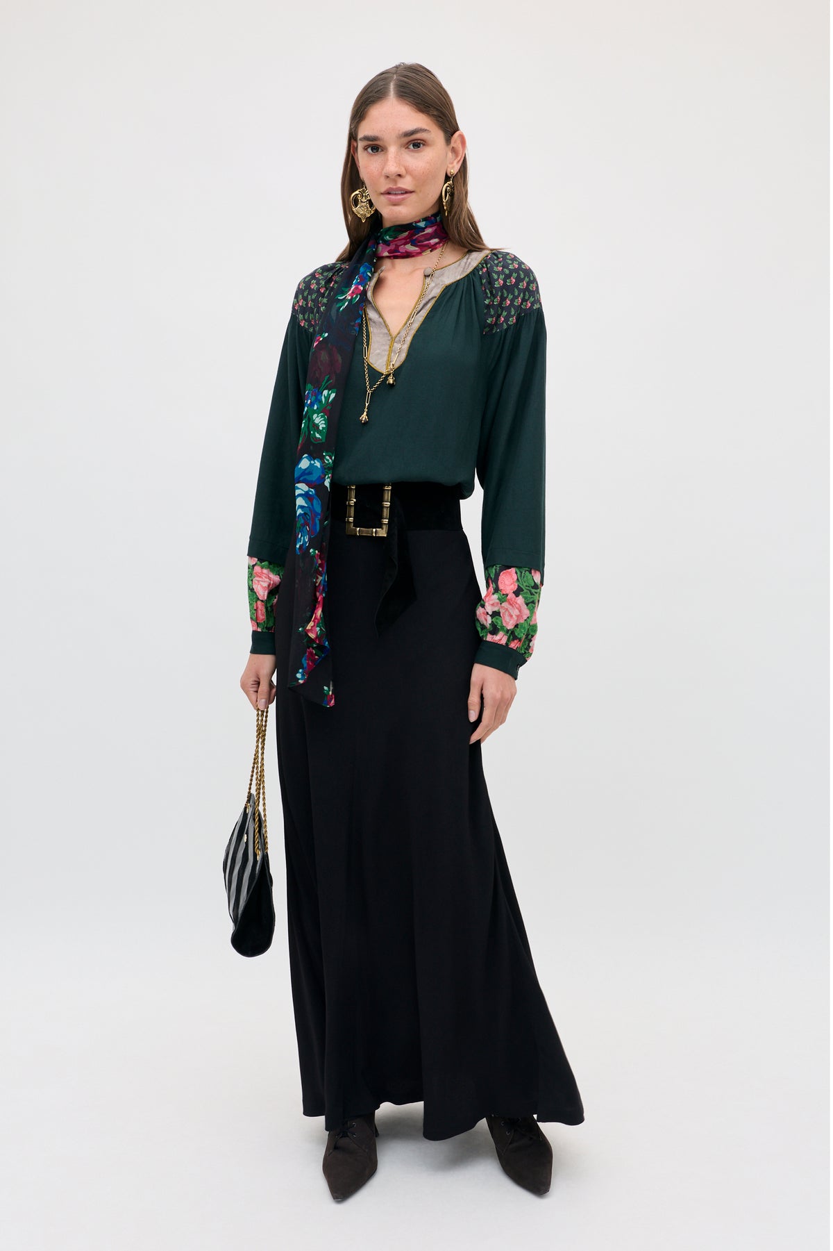 A woman wearing a Julien - Vintage Bouquet Black floral-print blouse, a long black skirt with a wide belt, and a colorful patterned scarf, holding a small black shoulder bag.