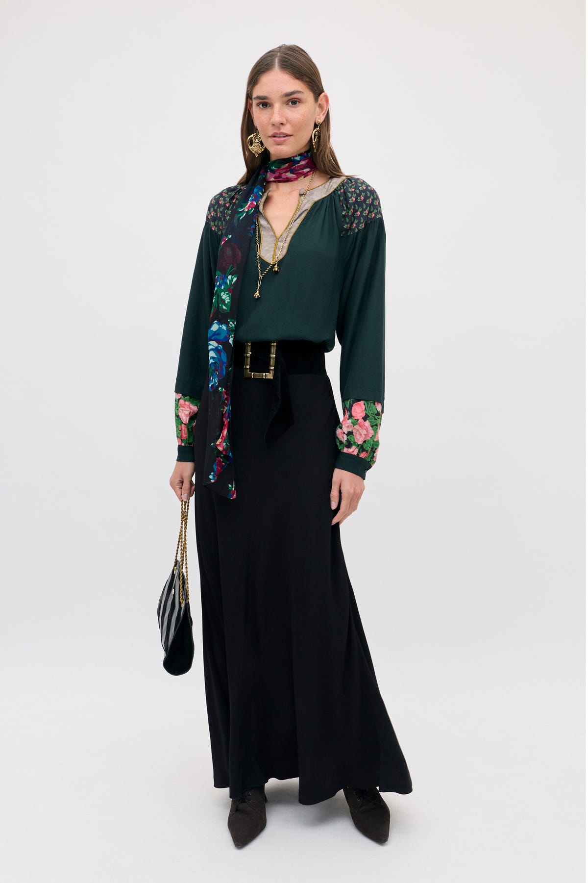 A woman wearing an Aide - Black top with floral cuffs, a matching scarf, a long black skirt, and a belt with a gold buckle, holds a small black handbag.