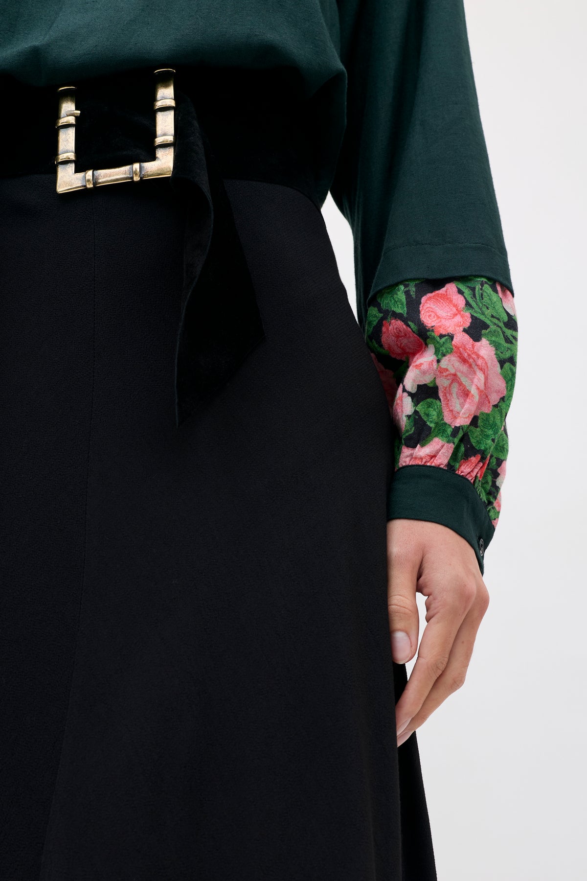 A person wearing an Aide - Black top with floral patterned cuffs, a black skirt, and a wide belt with a prominent rectangular gold buckle.