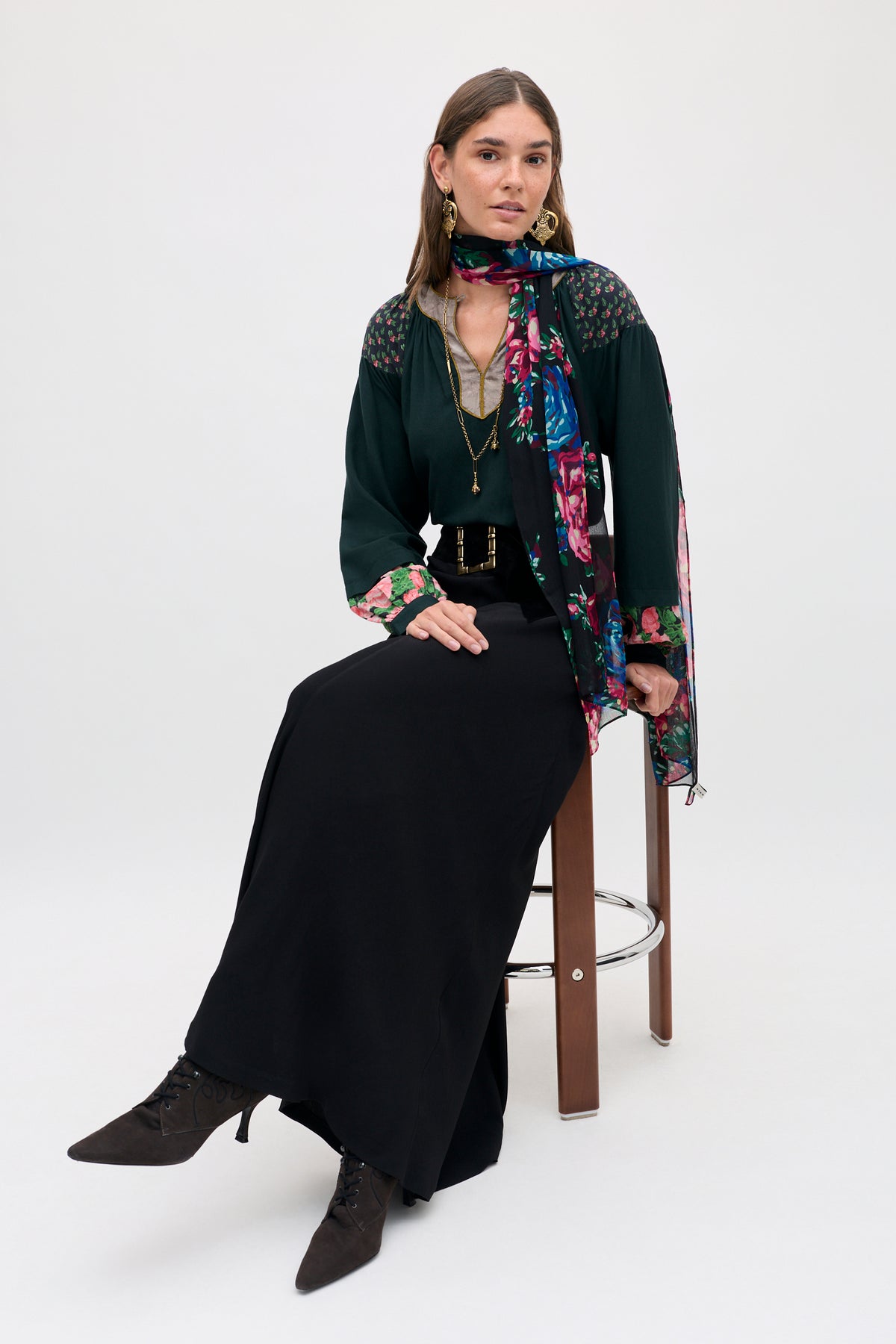 A woman sits on a stool wearing a Julien - Vintage Bouquet Black long-sleeved dark green top with a floral print scarf, a black maxi skirt, and lace-up pointed-toe boots.