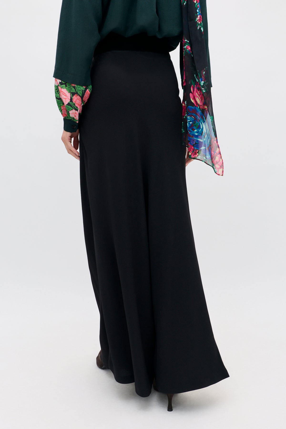 A person wearing an Aide - Black long skirt paired with a dark green long-sleeved shirt with floral patterned cuffs and a matching floral scarf.