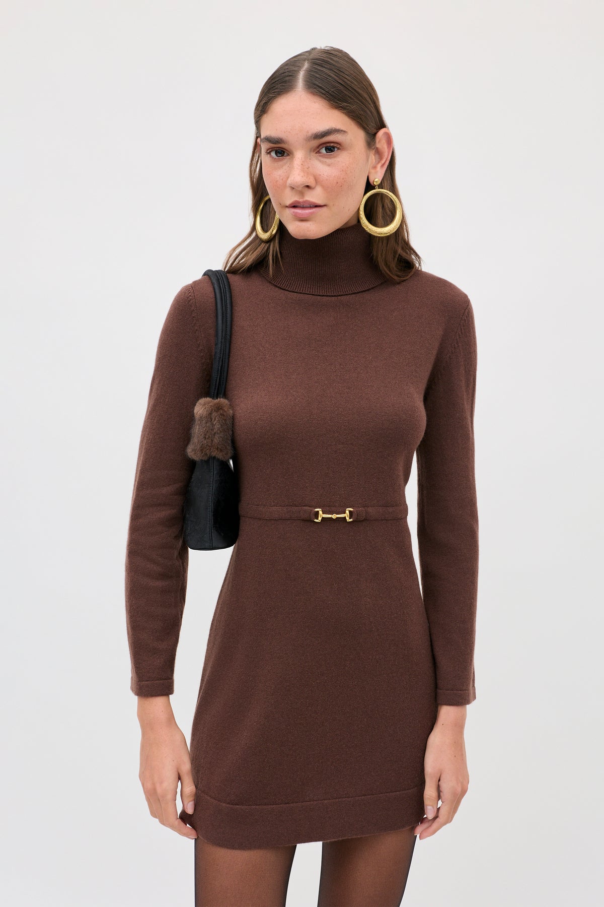 A woman wears the brown turtleneck Aly - Chocolate mini dress with a thin, gold-toned horsebit belt and carries a small black shoulder bag.