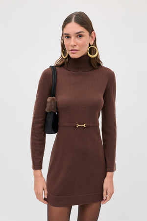 A woman wears the brown turtleneck Aly - Chocolate mini dress with a thin, gold-toned horsebit belt and carries a small black shoulder bag.