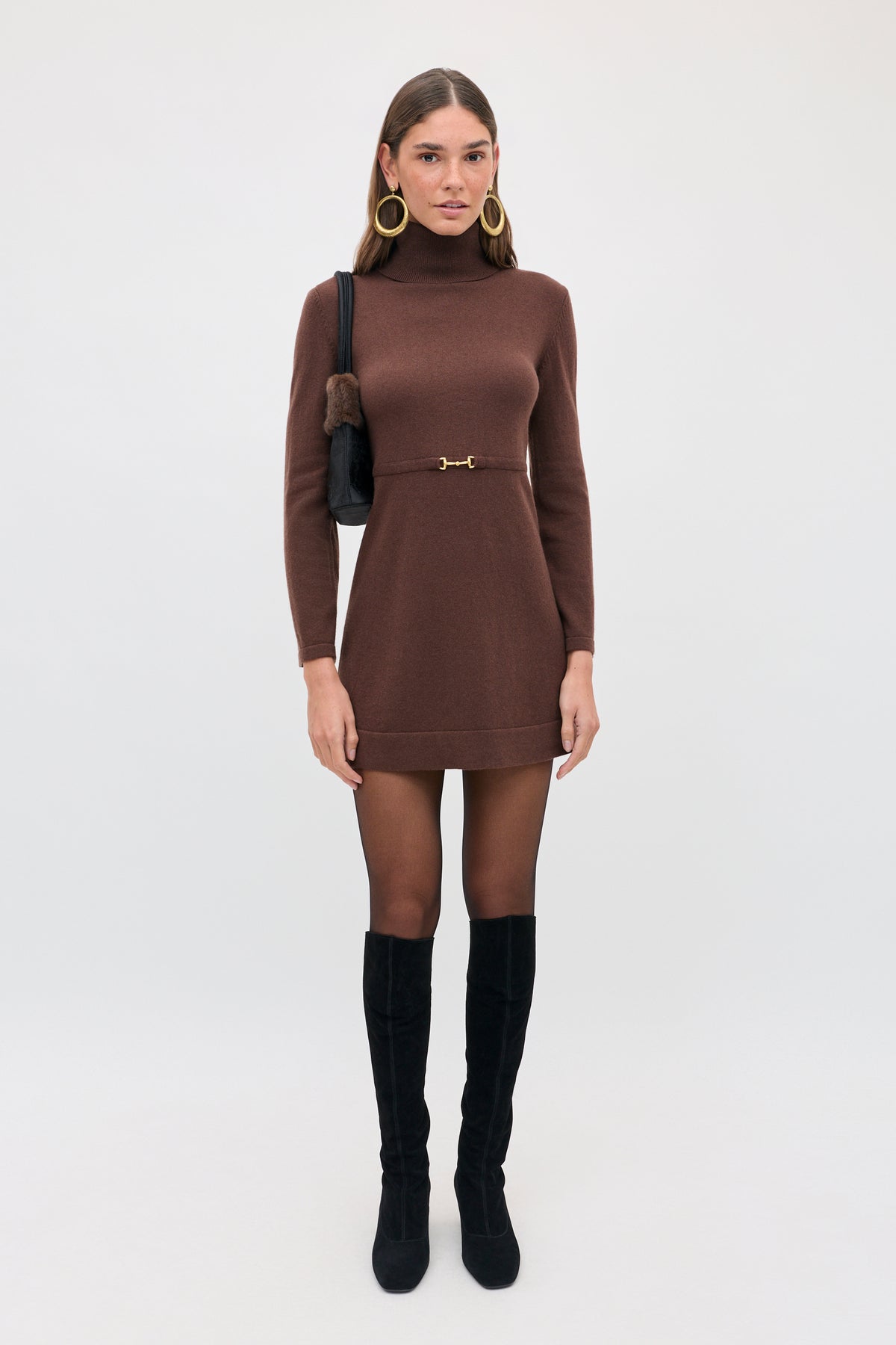 A woman wearing an Aly - Chocolate brown turtleneck mini dress with a gold waist detail, black tights, knee-high black boots, and a small black shoulder bag.