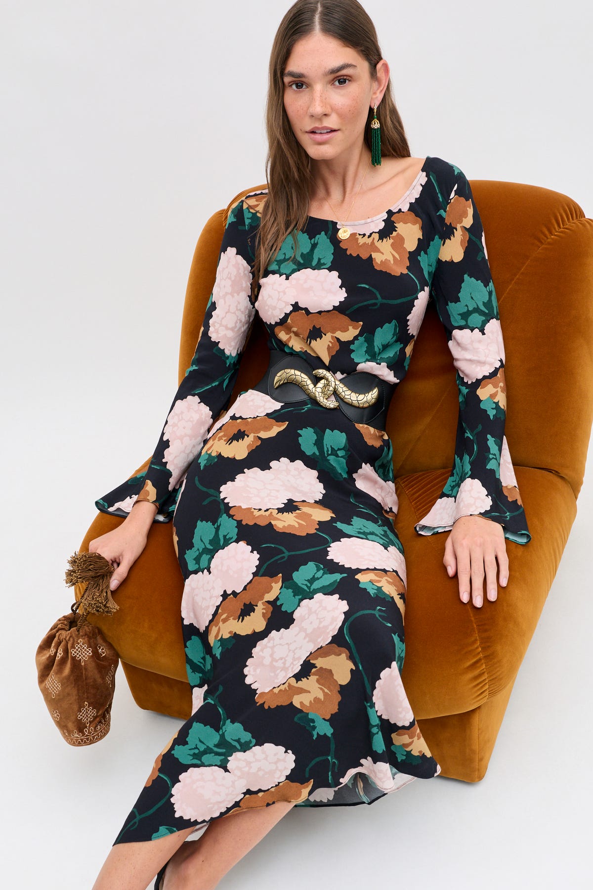 A woman wears the Amal - Hydrangea Floral Charcoal midi dress, featuring a long-sleeved dark charcoal hydrangea floral print, accented by a wide black belt with a gold snake buckle and a matching brown drawstring pouch.
