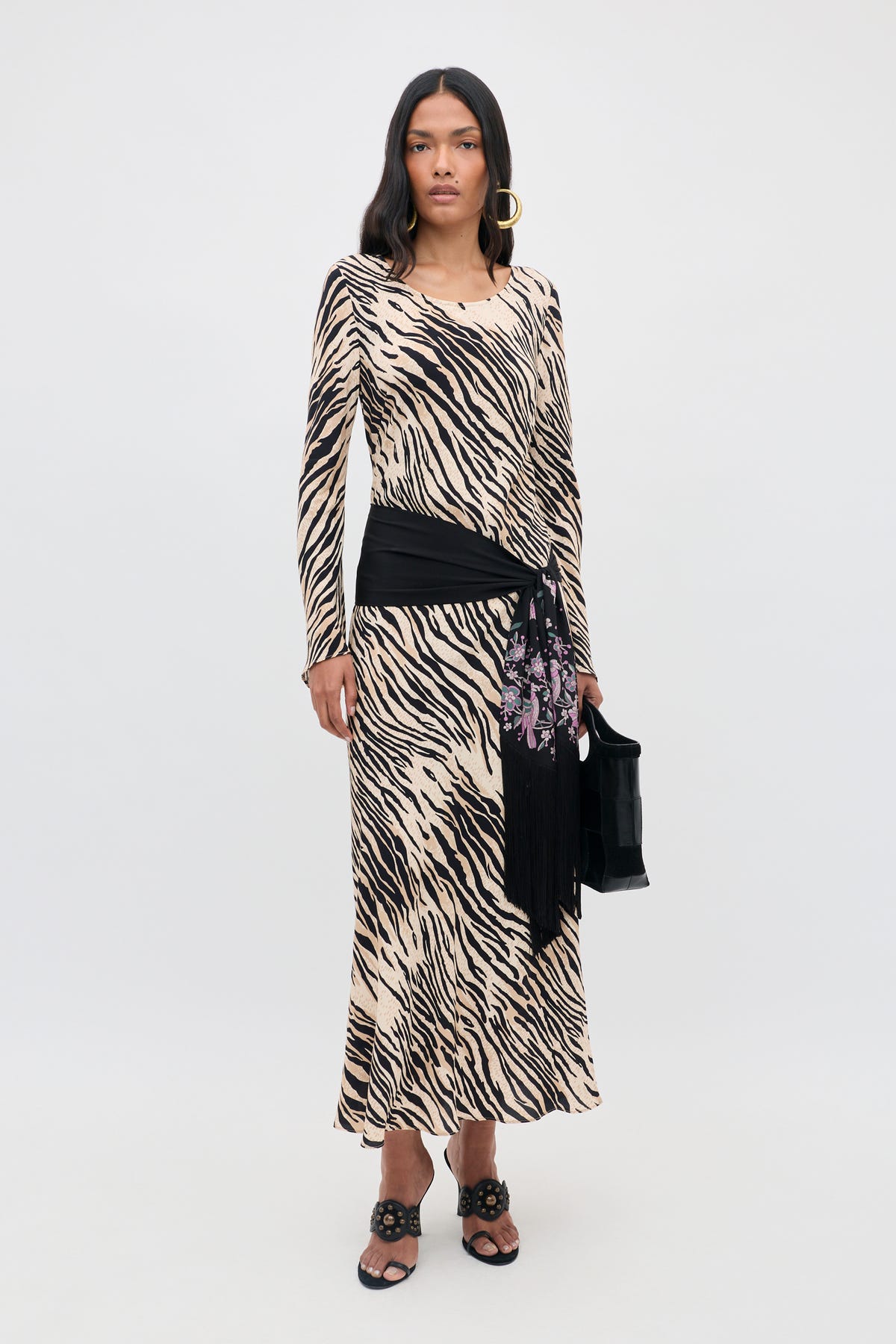 A woman wearing the Amal - Cooper Tiger Beige long-sleeve maxi dress with a black hip scarf, black sandals, and holding a black handbag stands against a plain white background.