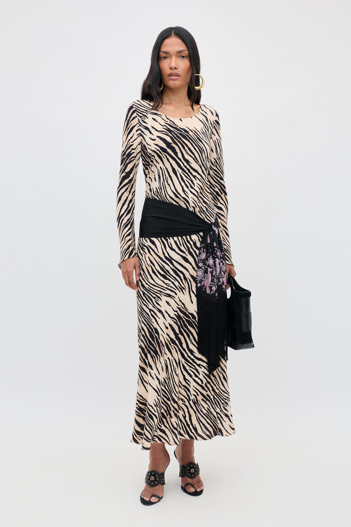 A woman wearing the Amal - Cooper Tiger Beige long-sleeve maxi dress with a black hip scarf, black sandals, and holding a black handbag stands against a plain white background.