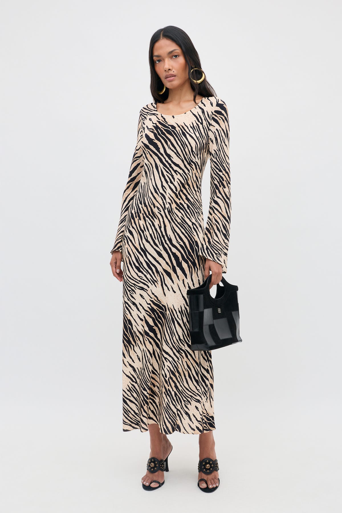 A woman wearing the Amal - Cooper Tiger Beige long-sleeved maxi dress, paired with black sandals and large hoop earrings, holds a small black handbag while standing against a plain light gray background.