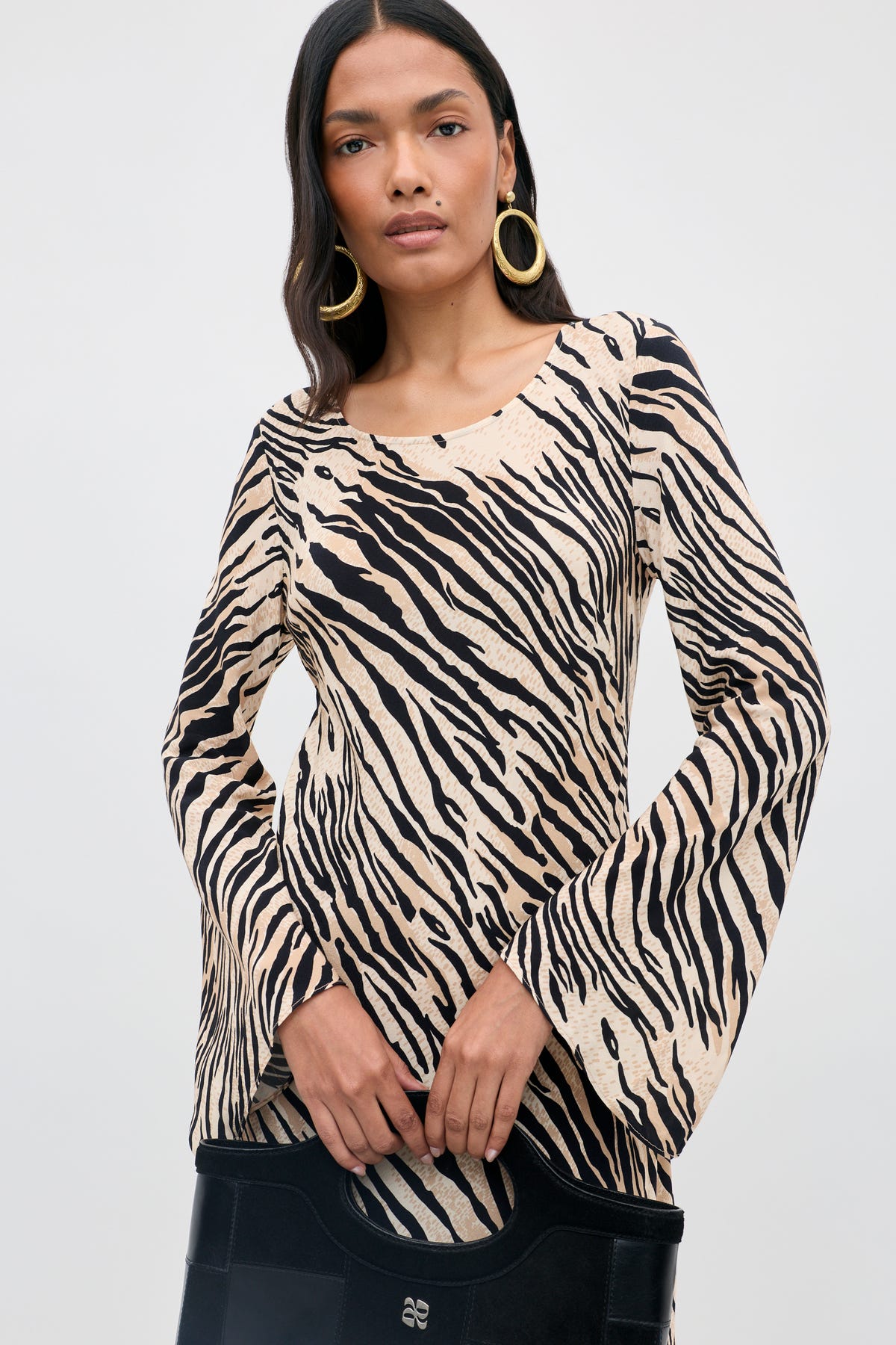 A woman stands against a plain, light background wearing the Amal - Cooper Tiger Beige long-sleeved zebra-print top, paired with a black bag and large gold hoop earrings, her straight dark hair framing her face.