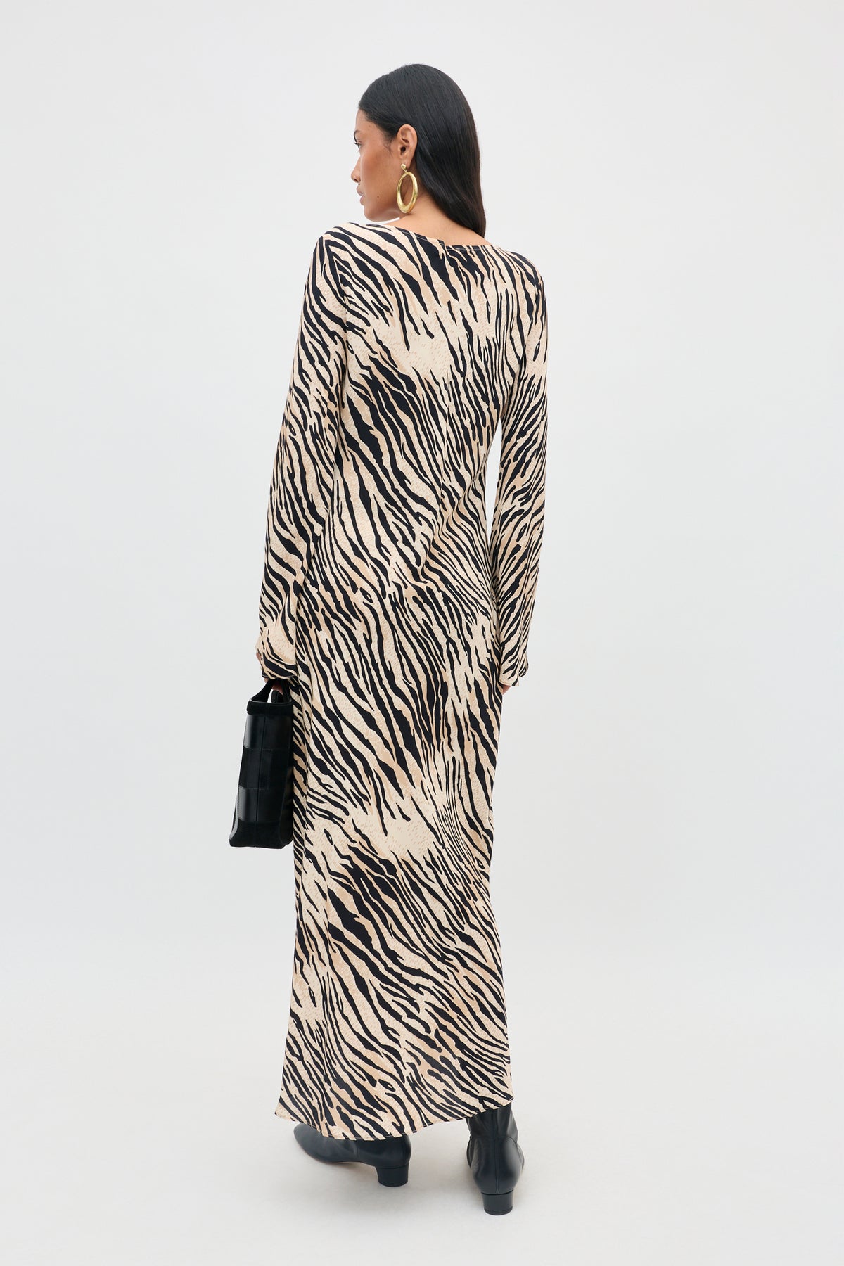 A person stands facing away, wearing the Amal - Cooper Tiger Beige long-sleeved, ankle-length dress with a bold black and beige zebra print. She pairs it with black ankle boots, large hoop earrings, and holds a black handbag.