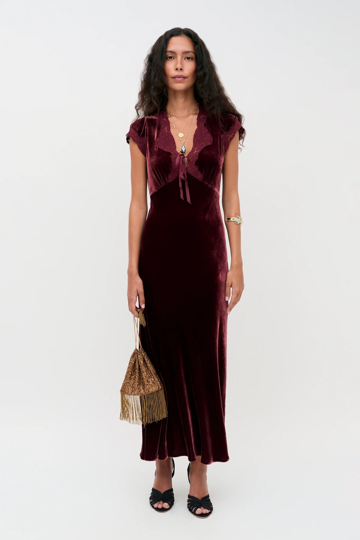 A person wearing the floor-length, deep red velvet Amari - Bordeaux gown with a lace-trimmed V-neckline, holding a gold beaded handbag.
