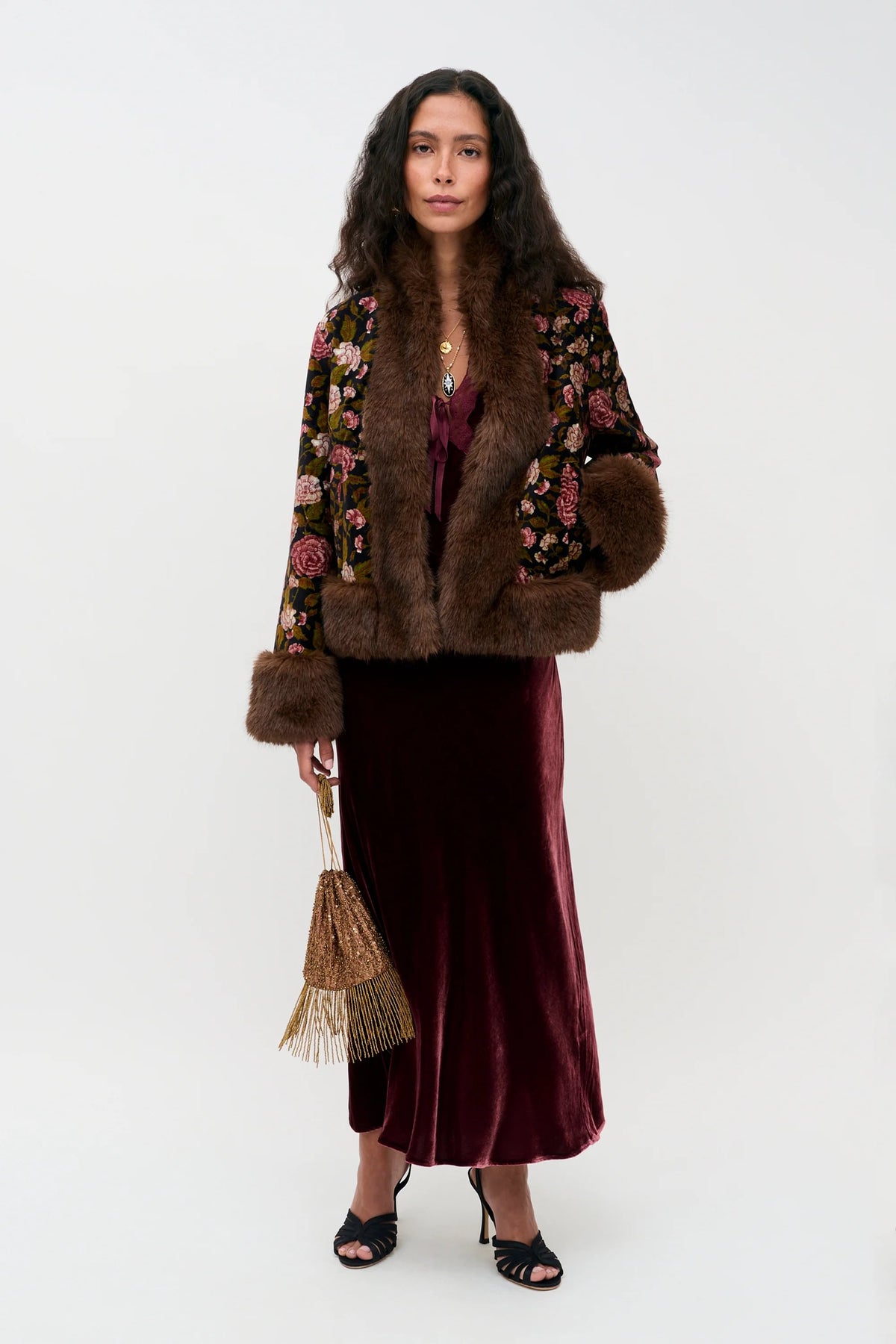 A person wearing the Amari - Bordeaux, featuring a floral print jacket with faux fur trim, a burgundy velvet midi dress, and black heeled sandals, while holding a small gold beaded bag.
