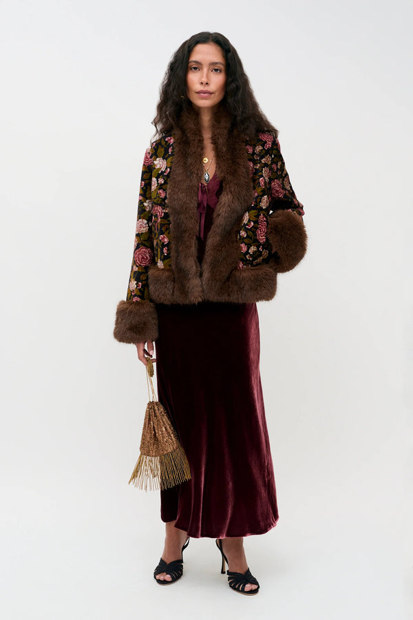 A person wearing the Amari - Bordeaux, featuring a floral print jacket with faux fur trim, a burgundy velvet midi dress, and black heeled sandals, while holding a small gold beaded bag.