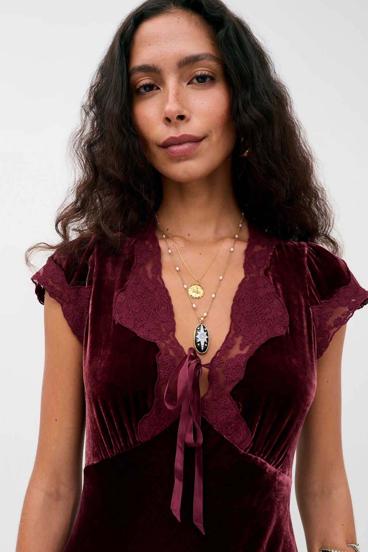 A woman wearing the Amari - Bordeaux, a dark red velvet V-neck dress with lace trim and layered gold necklaces.