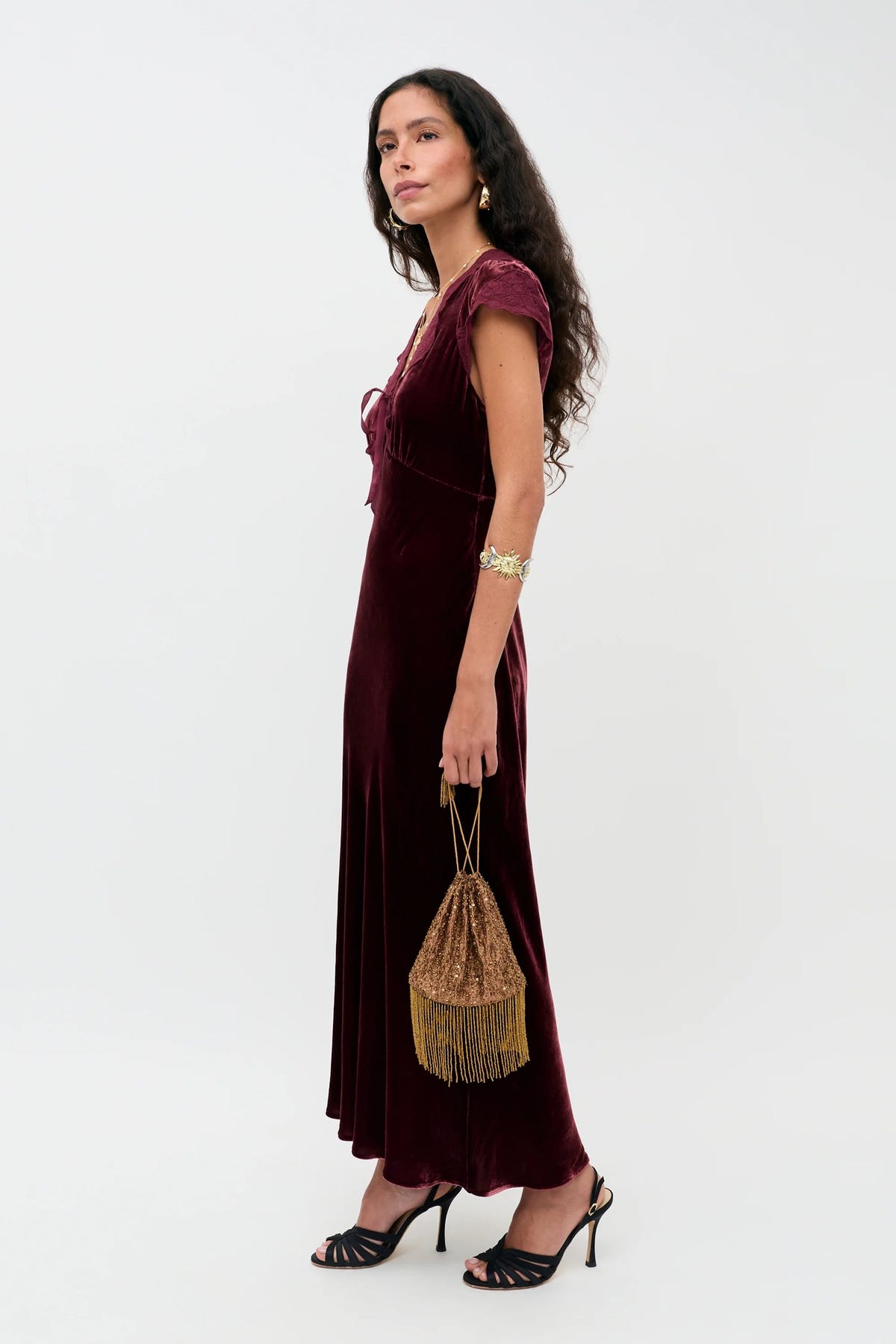 A woman wears the Amari - Bordeaux, a floor-length burgundy dress with black heeled sandals, while holding a small gold beaded bag with a fringe.