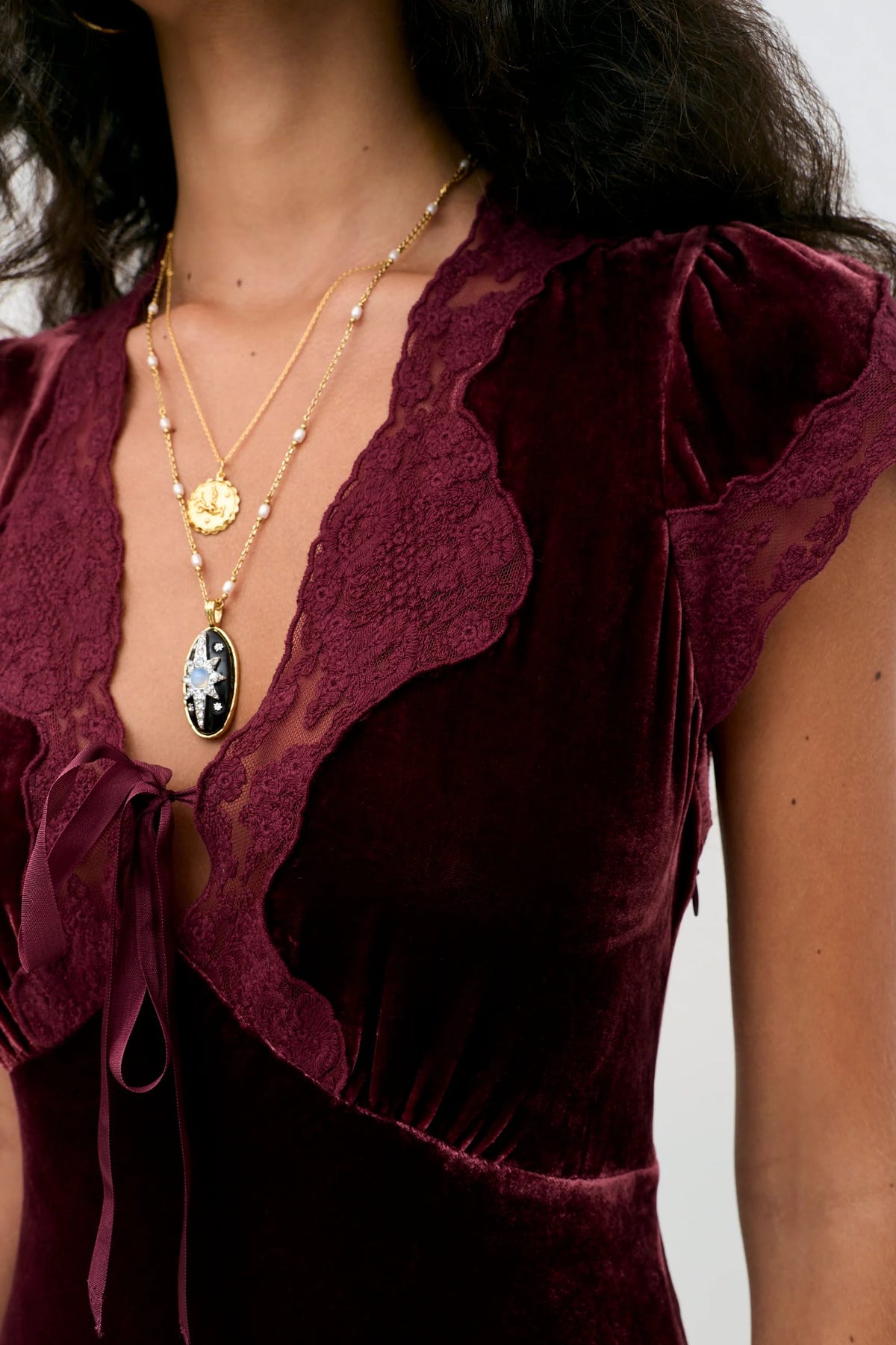A person wears the Amari - Bordeaux, a dark red velvet dress with lace trim and gold layered necklaces featuring an oval pendant.