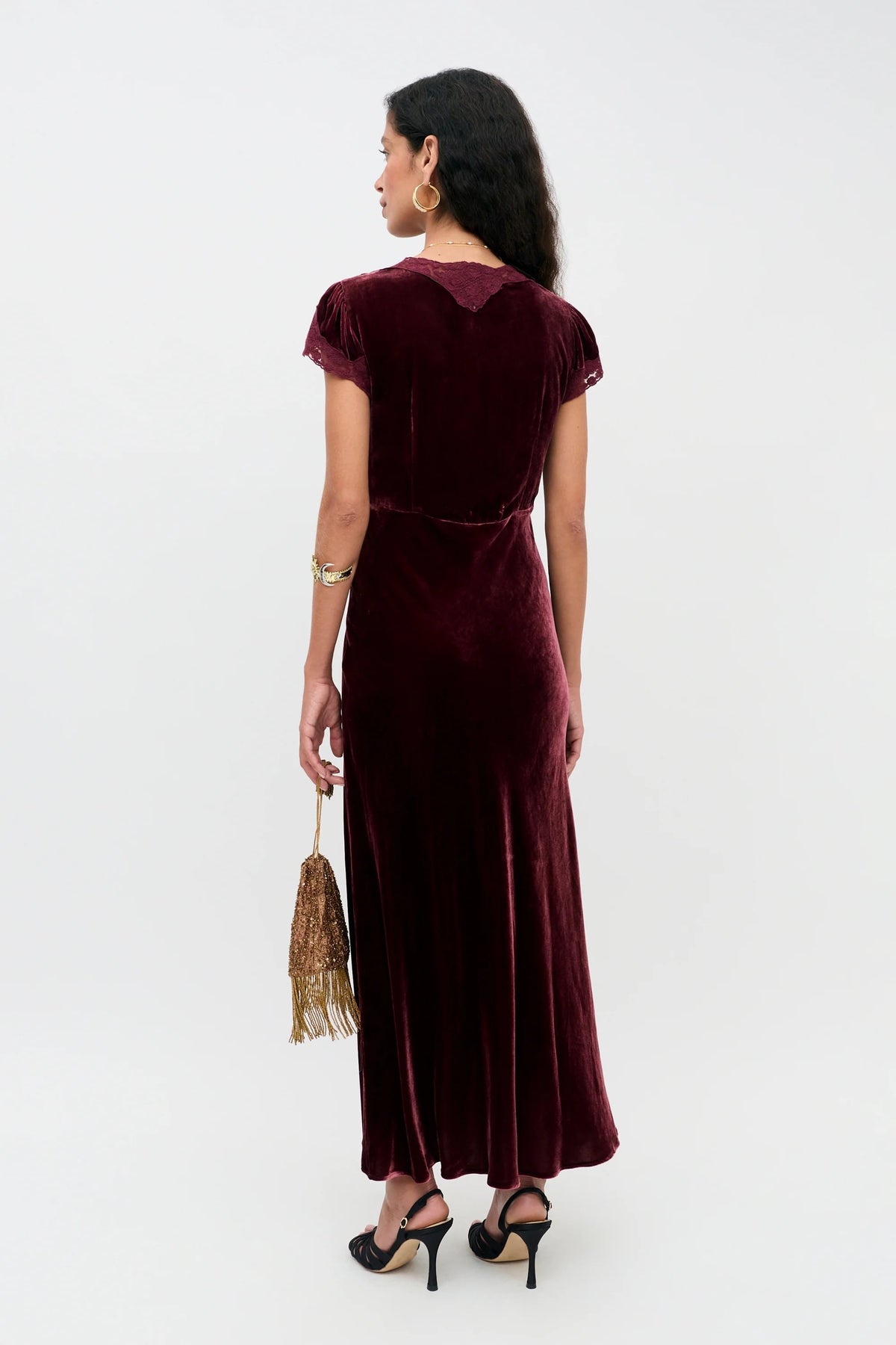 A woman standing with her back to the camera, wearing the Amari - Bordeaux floor-length dress with short sleeves and holding a small gold tasseled bag.