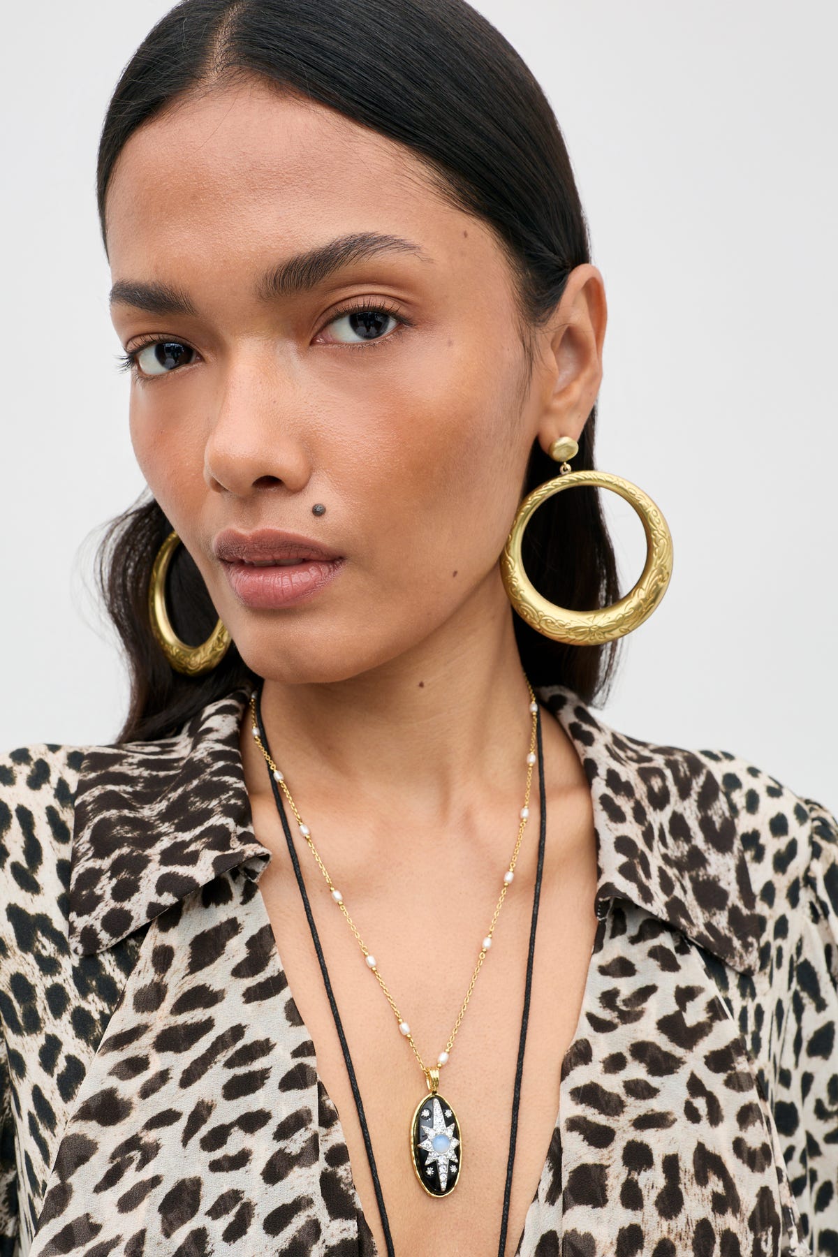 A woman with straight dark hair and a beauty mark above her lip gazes at the camera with a neutral expression, wearing the Andi - Black top paired with large gold hoop earrings and layered gold necklaces.