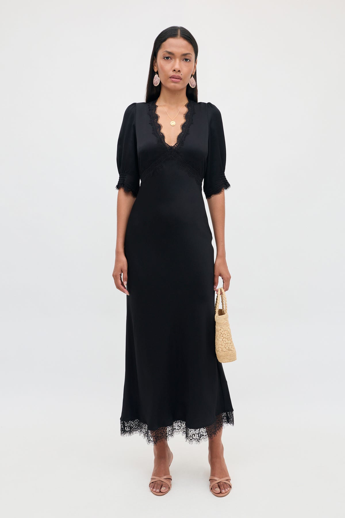 Clad in the Annina - Black dress, she showcases its v-neckline and short sleeves, accented by delicate lace trims at the hem. The bias cut of this ankle-length midi dress flatters her silhouette as she holds a small woven bag. Completing her look with sandals, statement earrings, and layered necklaces, she stands against a plain white background.