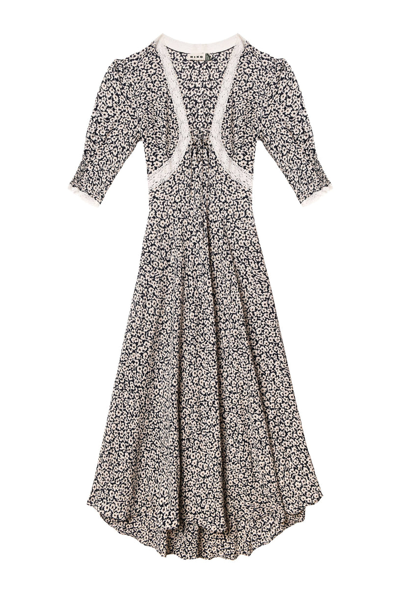 The Amina - English Daisy Deep Navy dress features a vintage-inspired navy blue design with white floral print, lace trim at the neckline and cuffs, short puff sleeves, a fitted waist, and a flared skirt.